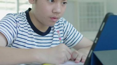 İyi görünümlü Asyalı çocuk evde bir sandalyeye otur ve cevapları bulmak için bir tablet kullanarak ev ödevini yapmak.