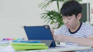 İyi görünümlü Asyalı çocuk evde bir sandalyeye otur ve cevapları bulmak için bir tablet kullanarak ev ödevini yapmak.