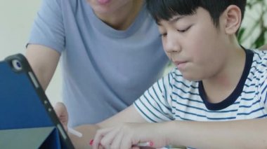 Asyalı anne ev ödevini yapmak için oğlunu kontrol ediyor. Oğlu raporu bulmak için tableti kullanarak ev ödevi yapıyor..