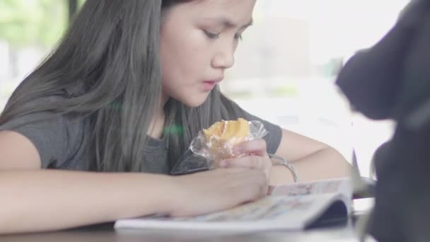 カジュアルな服を着たかわいいアジアの女の子が座って本を読んだり カフェでケーキを食べたりして楽しむ — ストック動画