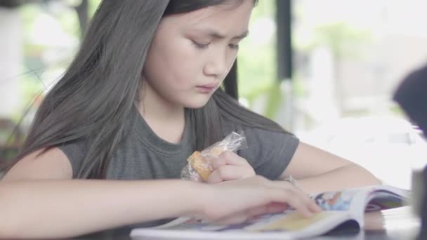可爱的亚洲小女孩穿着休闲的衣服坐在享受阅读书和吃一块蛋糕在咖啡馆 — 图库视频影像
