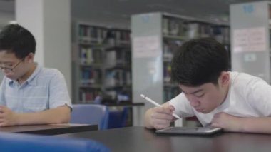 Asyalı ergenler kütüphanede tablet bilgisayar kullanıyorlar. Tablet bilgisayarına gülen yüzlü iki çocuk çiziyor..