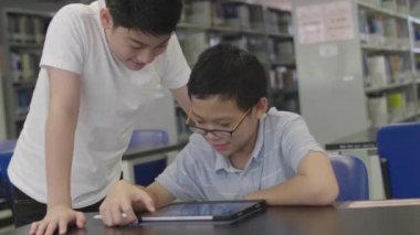Asyalı ergenler kütüphanede tablet bilgisayar kullanıyorlar. Tablet bilgisayarına gülen yüzlü iki çocuk çiziyor..