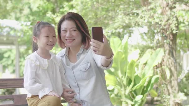 アジアの明るい母とともに彼女の娘は庭に座って スマートフォンで自撮り写真を与える — ストック動画