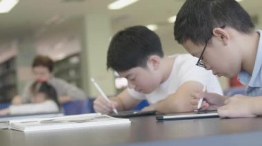 Asyalı ergenler kütüphanede tablet bilgisayar kullanıyorlar. Tablet bilgisayarına gülen yüzlü iki çocuk çiziyor..