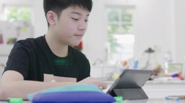 Asyalı genç öğrenci evde online çalışıyor. Çocuk öğretmenle çevrimiçi öğretim yoluyla iletişim kuruyor..