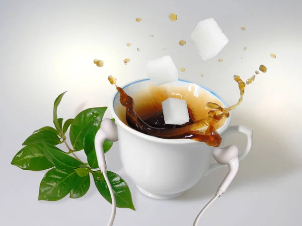 A white cup with white earphones put on it, splash of coffee, three  sugar cubes and green  coffee leaves.