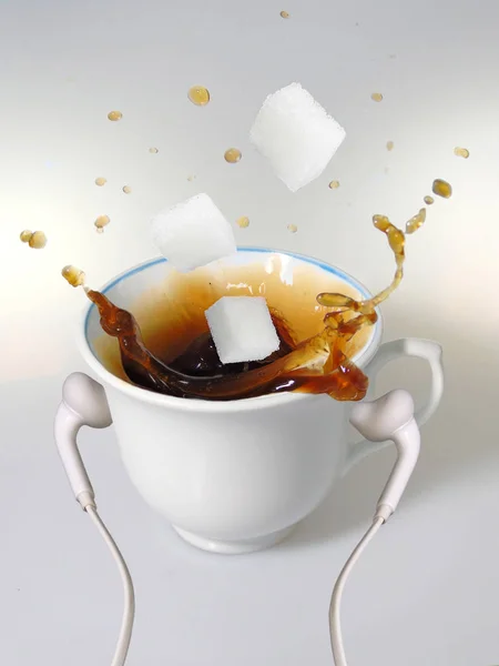 A white cup with white earphones put on it, splash of coffee, and three  sugar cubes.