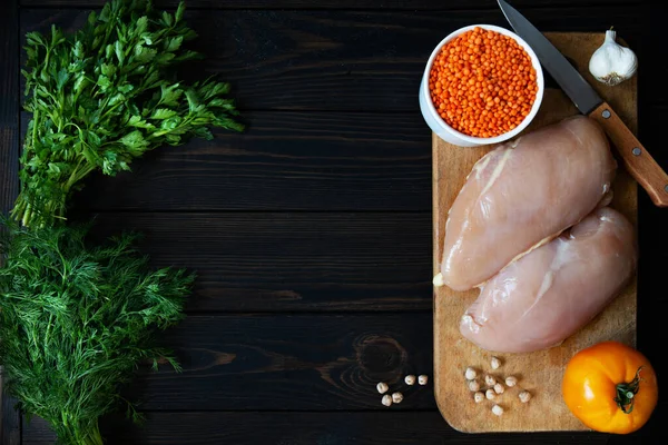 Fresh raw chicken fillet on a cutting board with herbs and vegetables on a dark background. Diet food top view with place for text.