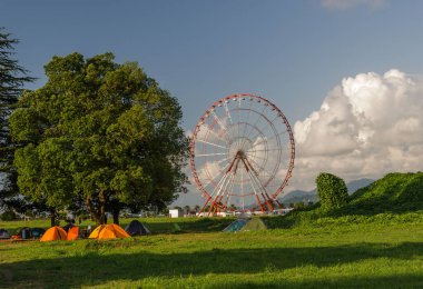 Batumi,Adjara,Georgia - July 06 2019, Batumi embankment, Ferris wheel in the rays of the setting sun, in the foreground on the green grass the tent of campers