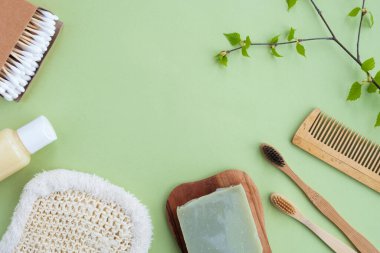 Yeşil arka planda plastiksiz banyo aksesuarları. Bambu diş fırçaları, şampuan şişesi, pamuk tomurcukları, tarak, doğal sabun, bez ve yapraklı dal. Üst görünüm, düz görünüm, kopyalama alanı