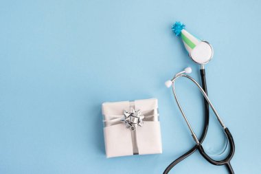 Şenlik şapkalı tıbbi steteskop ve açık mavi arka planda hediye. Doktorların günü, Dünya Sağlık Günü. Teşekkürler doktorlar ve hemşireler. Yaratıcı tıbbi kartpostal. Üst görünüm, düz görünüm, kopyalama alanı