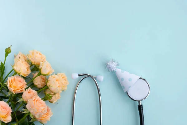 Şenlik şapkasında tıbbi steteskop ve açık mavi arka planda çiçekler. Doktorların günü, Dünya Sağlık Günü. Teşekkürler doktorlar ve hemşireler. Yaratıcı tıbbi geçmiş, kartpostal. Üst görünüm, düz görünüm, kopyalama alanı