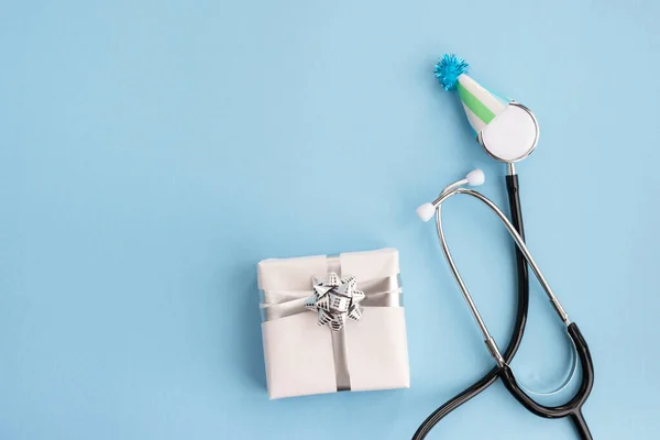 Şenlik şapkalı tıbbi steteskop ve açık mavi arka planda hediye. Doktorların günü, Dünya Sağlık Günü. Teşekkürler doktorlar ve hemşireler. Yaratıcı tıbbi kartpostal. Üst görünüm, düz görünüm, kopyalama alanı