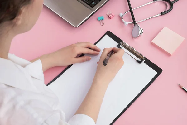 Pembe masada tıbbi ekipman ve dizüstü bilgisayarı olan bir doktor. Kadın doktor panoya yazı yazıyor. Sağlık ve tıp konsepti. Üst manzara, düz uzanma