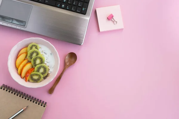Healthy food on working table. Muesli with natural yogurt, peach and kiwi on light pink background. Snack at workplace, food in the office. Top view, flat lay, copy space