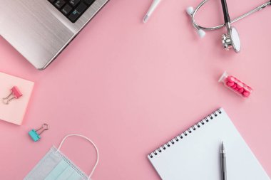 Doktor işyeri ve tıbbi ekipmanlar pembe arka planda. Stetoskop, laptop, koruyucu yüz maskesi, termometre, haplar, not defteri ve kalem. Üst görünüm, düz görünüm, çerçeve, kopyalama alanı