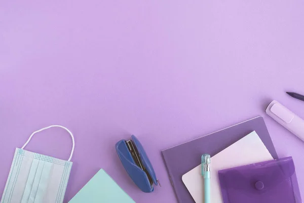 Okula geri dön, koronavirüs salgını konsepti. Pastel mor arka planda kırtasiye malzemeleri ve tıbbi yüz maskesi. Üst görünüm, düz görünüm, kopyalama alanı