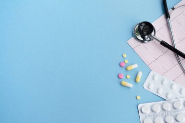 Tıbbi steteskop, kardiyogram ve açık mavi arka planda haplar. Kardiyoloji konsepti. Boşluğu kopyala, üst görünüm