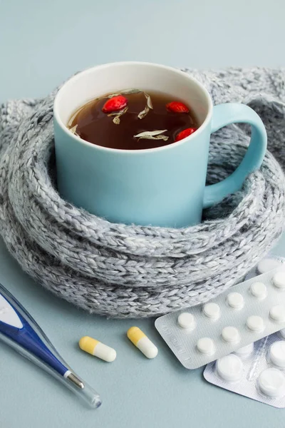 Cold and flu season. Mug of hot tea with herbs and rosehips, thermometer and pills on light blue background. Treatment and prevention of the virus. Vertical