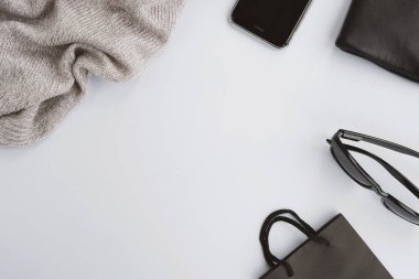 Gri arka planda moda aksesuarları. Süveter, el çantası, akıllı telefon, güneş gözlüğü ve kese kağıdı. Alışveriş, Kara Cuma, sonbahar ya da kış mevsimi konsepti. Üst görünüm, düz görünüm, kopyalama alanı