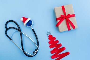 Kırmızı Noel Baba şapkasında tıbbi steteskop hediye kutusu ve açık mavi arka planda kırmızı kurdeleden yapılmış Noel ağacı. Noel ve Yeni Yıl konsepti. Yaratıcı tıbbi kış geçmişi, kartpostallar. Üst görünüm