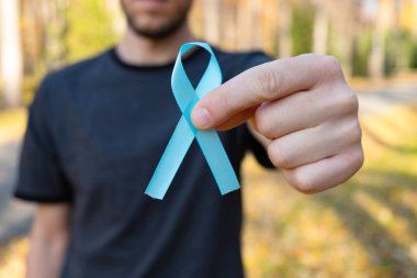 İnsan elleri sonbahar ormanının arka planında mavi prostat kanseri farkındalığı kurdelesini tutuyor. Tıp ve sağlık konsepti, erkek sağlığı. Kapat.