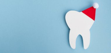 Açık mavi arka planda kırmızı Noel Baba şapkalı dekoratif diş. Noel ve Yeni Yıl konsepti. Diş hekimliği için yaratıcı tıbbi kış kartpostalı. Üst görünüm, pankart, kopyalama alanı