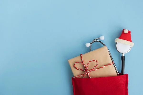 Noel Baba 'nın kırmızı çantasında hediye kutusuyla birlikte şapka takan tıbbi steteskop. Noel ve Yeni Yıl konsepti. Yaratıcı tıbbi kış tebrik kartı. Üst görünüm, düz görünüm, kopyalama alanı