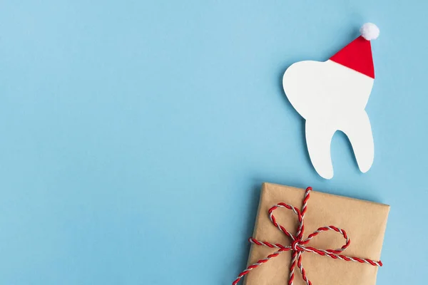 Kırmızı Noel Baba şapkalı dekoratif diş ve açık mavi arka planda hediye. Noel ve Yeni Yıl. Diş hekimliği için yaratıcı tıbbi kış kartpostalı. Üst görünüm, pankart, kopyalama alanı