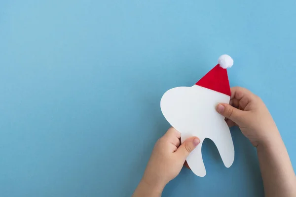 Kırmızı Noel Baba şapkasıyla açık mavi arka planda sevimli bir diş tutan küçük çocuk. Noel ve Yeni Yıl. Diş hekimliği için yaratıcı tıbbi kış kartpostalı. Üst görünüm, kopyalama alanı, düz uzanma