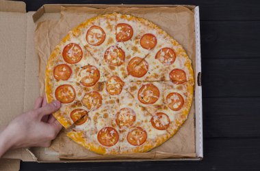 Peynirli pizza ve siyah ahşap arka planda bir kutuda domates. Genç bir adam bir dilim pizza alır. Reklam pizzacıları