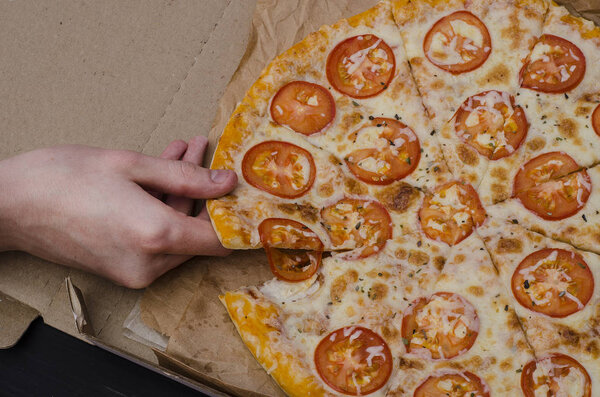 Pizza with cheese and tomatoes in a box on a black wooden background. A young man takes a slice of pizza. Advertising pizzerias, cafes.