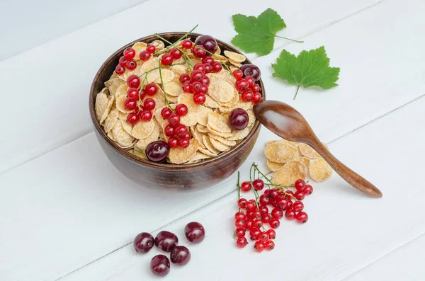 Fresh cornflakes with currants and cherries in a round bowl on a white wooden background.