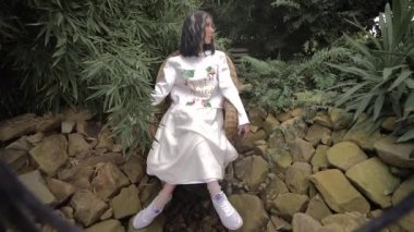 beyaz giysiler içinde esmer tropik bitkiler ile çevrili, taşlar üzerinde bir hasır sandalyede oturuyor. Kamera yukarıdan aşağıya doğru yönlendirilir. Saçta beyaz iplikçikler