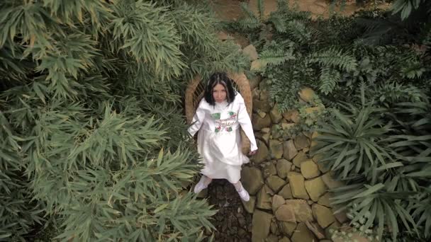 bruneta v bílých šatech sedí na proutěném křesle na kamenech, obklopeném tropickými rostlinami. Kamera směřuje shora dolů. Bílé prameny ve vlasech