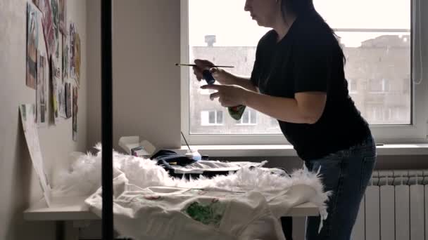 黒いTシャツとジーンズのブルネットは 窓の近くにブルテリアのイラストをジャケットに描きます 白い壁にはたくさんの絵がある — ストック動画