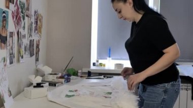 siyah bir t-shirt ve kot pencerenin yanındaki ceket tüyleri üzerinde çalışıyor esmer. Beyaz duvarda bir sürü çizim var..