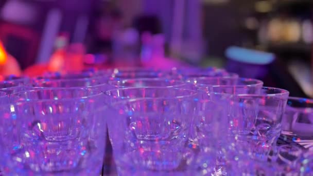 在酒吧柜台的一个黑暗的房间里 许多玻璃杯空玻璃是彼此的 背景中有五颜六色的灯光 — 图库视频影像