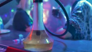 Leopar ceketli bir adam ve deri beregiymiş bir sarışın arka planda, mavi ışıklı bir odada konuşuyorlar. Bir cam nargile şişesinde, su gurgles, sarı ışık ile aydınlatılmış