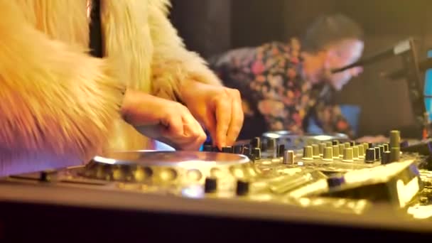 白い毛皮のコートを着たDjと胸の十字架がDjコンソールをコントロールします バックグラウンドの男は他の人とコミュニケーションをとります 部屋は黄色い光で照らされている — ストック動画