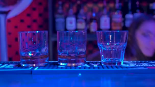 女の子のバーテンダーは飲み物をグラスにアイスキューブを注いだ バックグラウンドで アルコールのボトルとショーケース 部屋は青とピンクのライトで照らされています — ストック動画