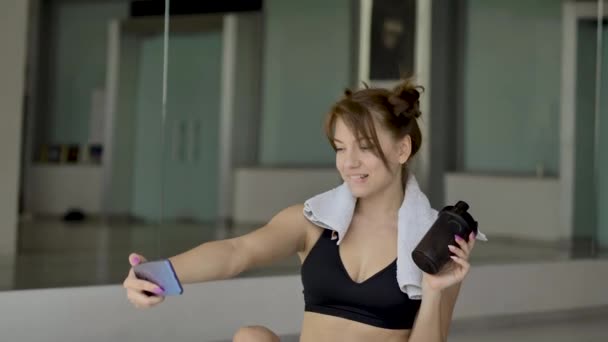 스포츠웨어의 스포티 소녀는 체육관에서 바닥에 앉아있는 전화로 자신을 촬영하고 있습니다 — 비디오