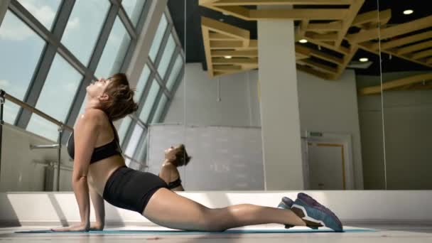 身着黑色运动服的白种人布鲁内特在地板上做伸展运动 在背景中 她在镜子里的倒影 一个彩色的玻璃窗和一台芭蕾舞机 — 图库视频影像