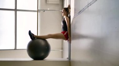 Spor giyim kafkas esmer beyaz salonda bir jimnastik topu ile egzersiz yapar, arka planda bir vitray pencere ve bir balerin için bir makine. Çok sıradışı bir kompozisyon
