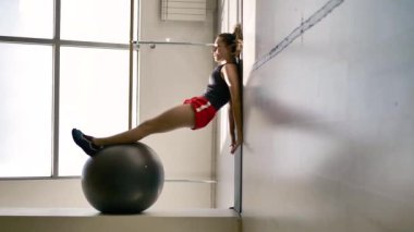 Spor giyim kafkas esmer beyaz salonda bir jimnastik topu ile egzersiz yapar, arka planda bir vitray pencere ve bir balerin için bir makine. Çok sıradışı bir kompozisyon