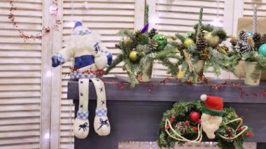 Kalın sakallı ve şapkalı mavi-beyaz bir Noel Baba bebeği, şöminenin üzerinde oturur, saksılarda Noel ağaçlarıyla süslenmiş, topları ve konileri, kırmızı oyuncaklı bir çelenk.