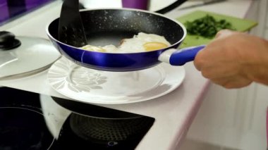 Kadın elleri omlet ve tabaklardan gıda ayırmak için çalışan bir kürek ile mavi bir tavada tutun, arka planda bir kesme tahtası, beyaz bir tabak ve bir kapak üzerinde yeşillik doğranmış.