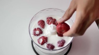 Yakın çekim, kadın elleri beyaz bir masa üzerinde duran bir blender için şeffaf bir fincan süt çilek koymak, üst görünümü.