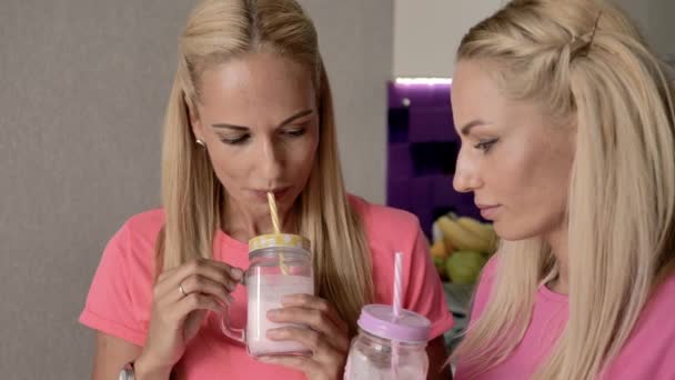 ブロンドの双子の姉妹は チューブ付きのガラスからミルクセーキを飲み ピンクの服を着て お互いに話し合い 良い気分です — ストック動画
